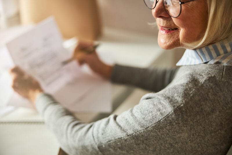 An elderly woman smiling while reviewing documents at a desk.