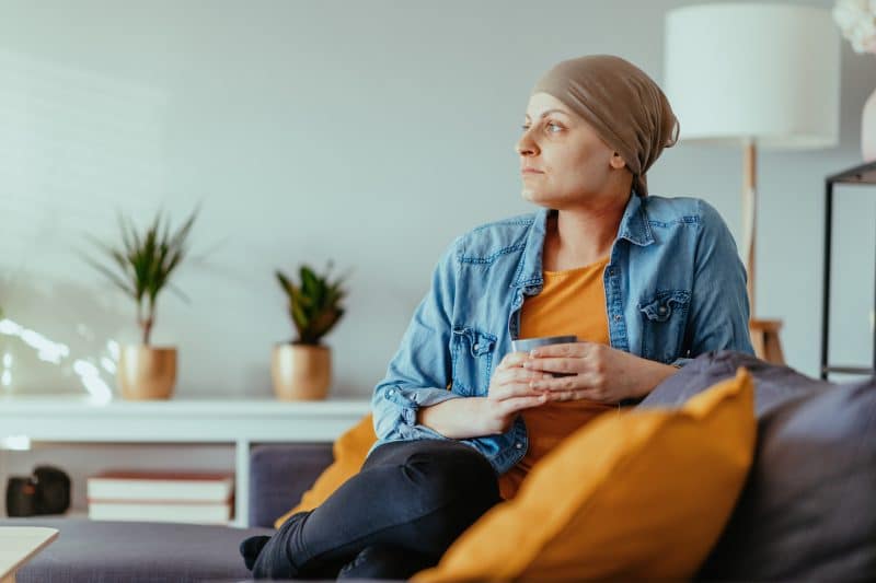 A woman undergoing cancer treatment sits on a couch, holding a cup.