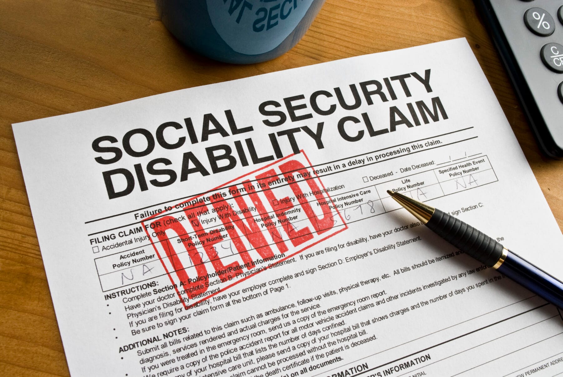 A Social Security Disability Claim form with a large red "DENIED" stamp across it, accompanied by a pen and a calculator on a wooden desk.