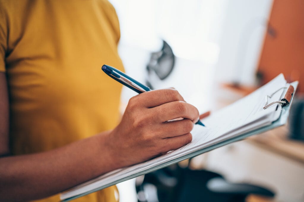 A person wearing a yellow shirt holds a clipboard and writes with a blue pen.