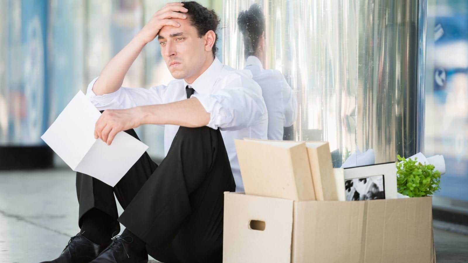 Man sitting on the ground in front of a building looking stressed, with a box of belongings next to him.