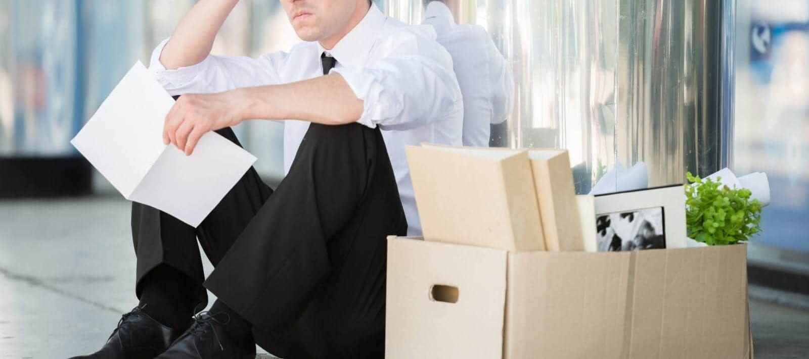 Man sitting on the ground in front of a building looking stressed, with a box of belongings next to him.