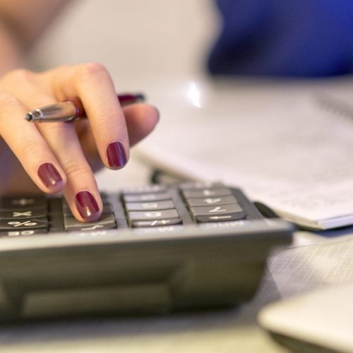 A close-up of a woman's hand using a calculator while holding a pen, with a notebook visible in the background.