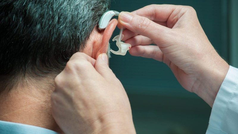 Close-up of a person being fitted with a behind-the-ear hearing aid by a medical professional.
