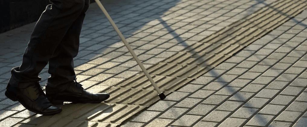 A blind man walking on pavement with a white cane.