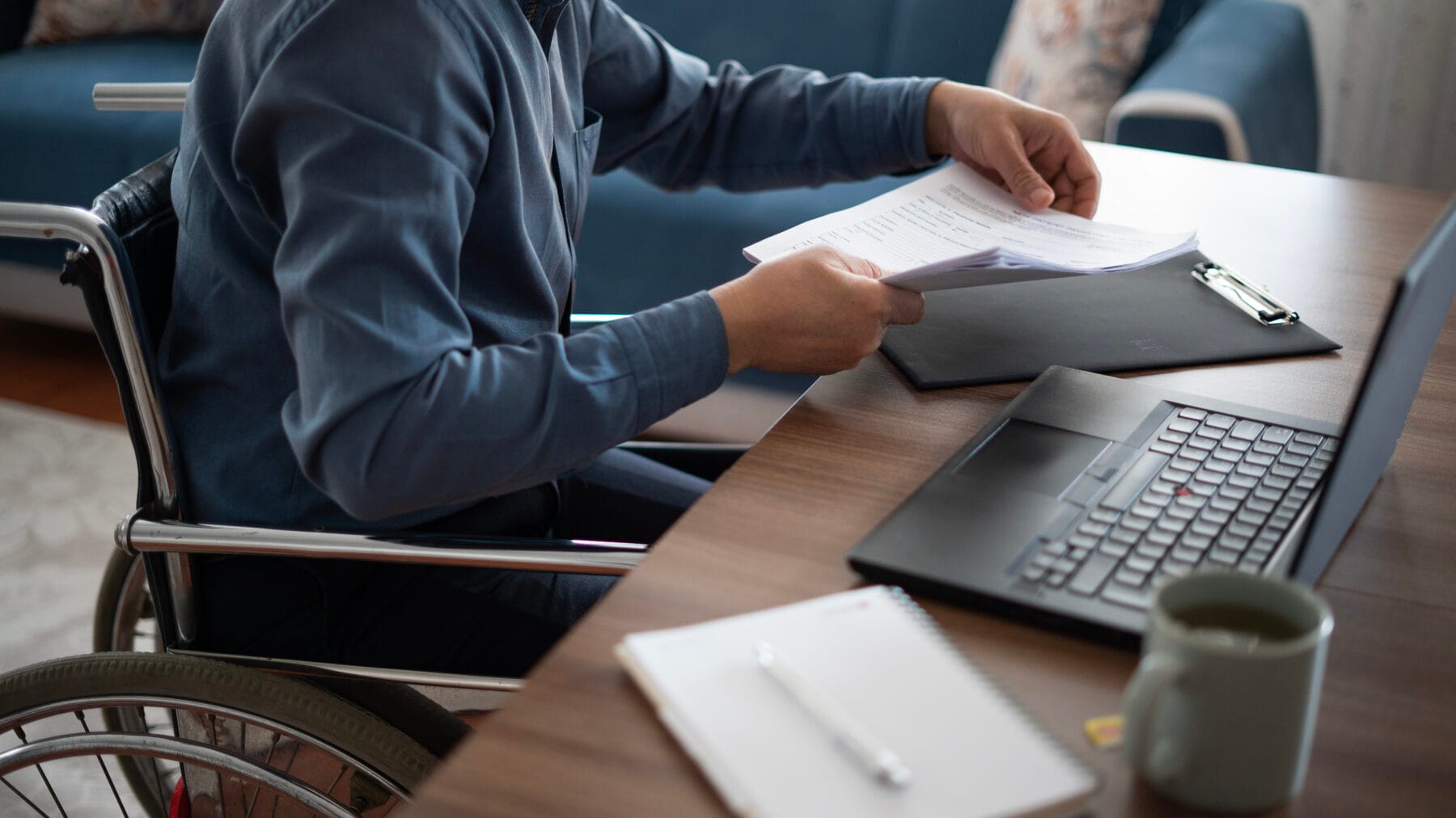 A person in a wheelchair is holding paperwork at a desk with a laptop, clipboard, notebook, and coffee mug.